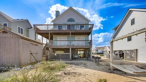 A multi-story beachfront house with a balcony, a covered porch, and a sandy yard.