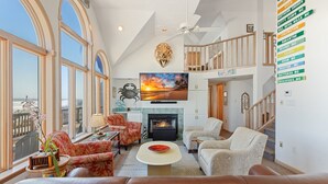 A spacious living room with a fireplace, comfortable seating, a large TV, and a view of the ocean through large windows. - Carpe Diem Corolla OBX  Pool HotTub (Corolla)