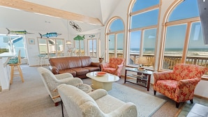A spacious living room with a large window overlooking the beach, a brown leather sofa, patterned armchairs, and a coffee table. - Carpe Diem Corolla OBX  Pool HotTub (Corolla)