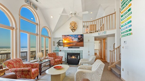 A spacious living room with large windows overlooking the beach, a fireplace, and a television mounted on the wall. - Carpe Diem Corolla OBX  Pool HotTub (Corolla)