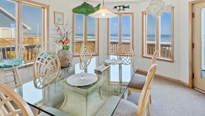 A dining area with a glass table, white chairs, and a view of the ocean through large windows. - Carpe Diem Corolla OBX  Pool HotTub (Corolla)