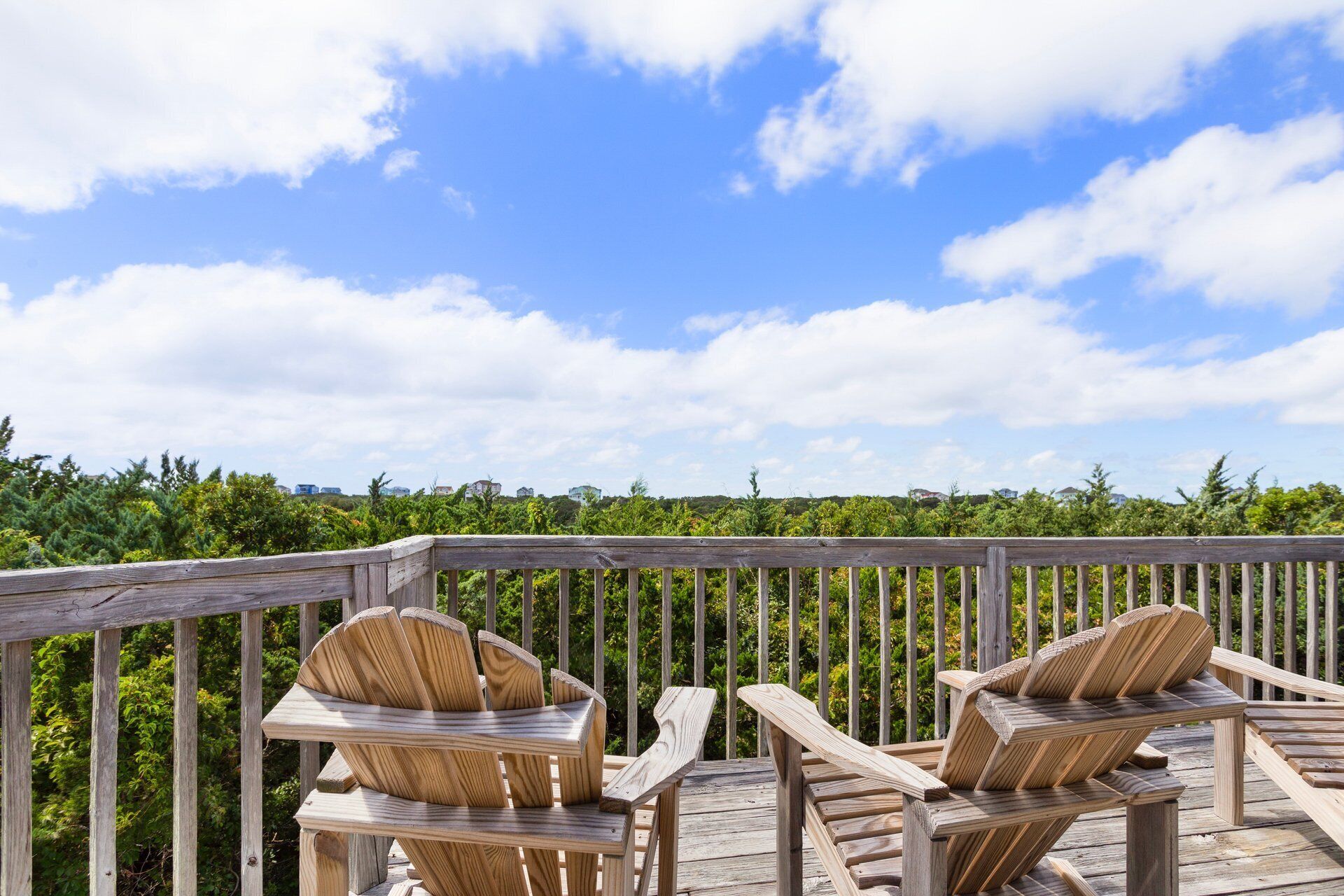 Two wooden Adirondack chairs on a wooden deck with a view of a forested landscape and a clear sky.