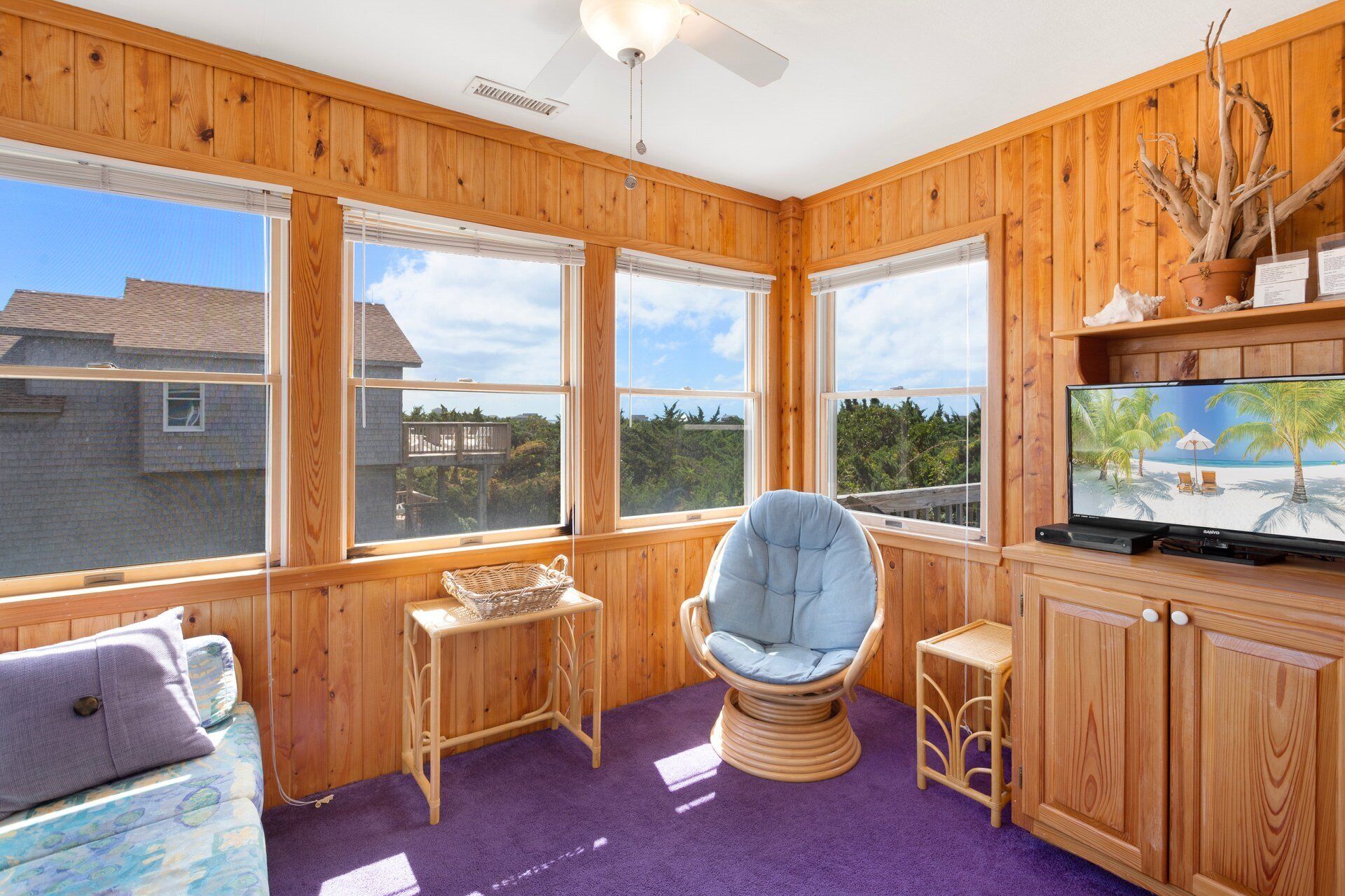 A room with wooden walls, large windows, a ceiling fan, a television, a wicker chair, a small table, and a purple carpet.