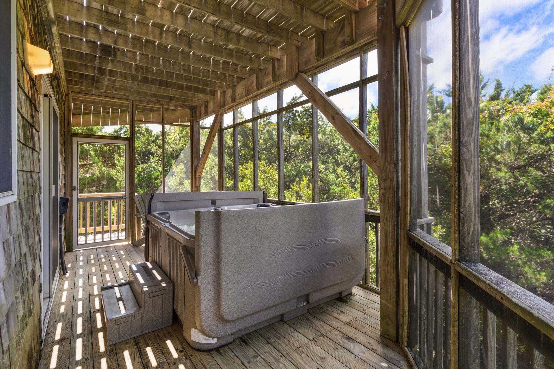 A wooden deck with a hot tub and a seating area, surrounded by large windows offering a view of the forest.