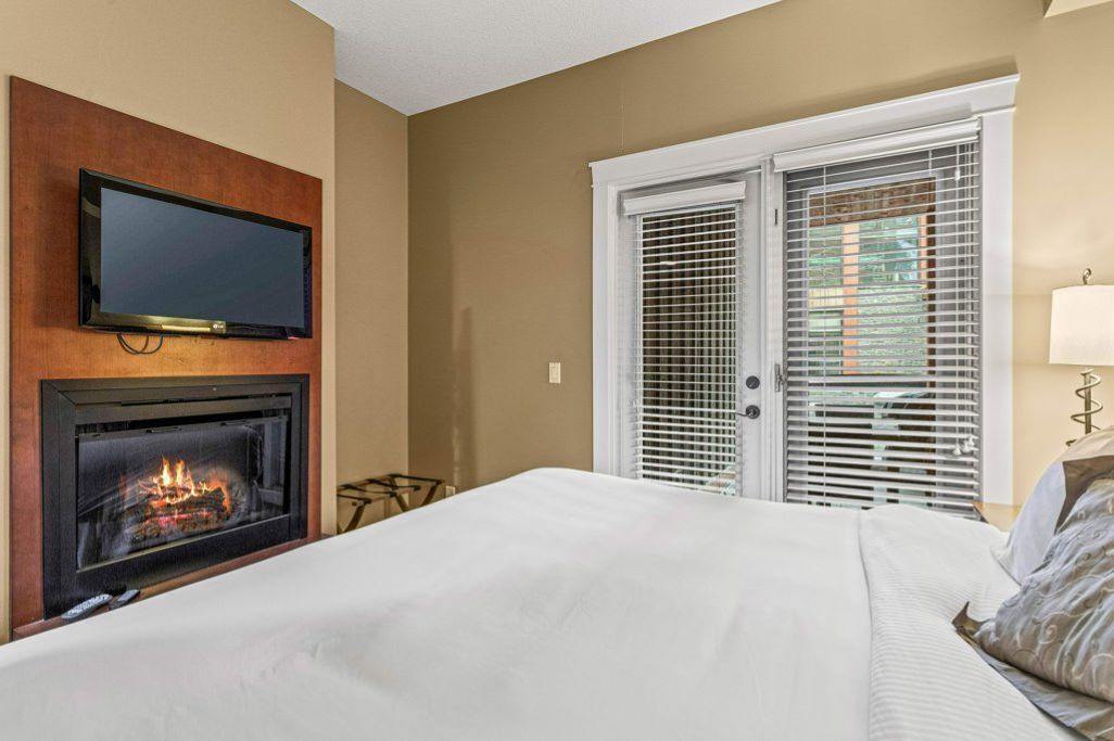 Ein Hotelzimmer mit Kamin, einem darüber montierten Fernseher, einem Bett mit weißer Bettwäsche und einem Fenster mit Jalousien.