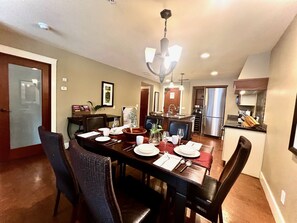 A dining area with a long table set for four, surrounded by chairs. A chandelier hangs above, and the room has a kitchen in the background. - A unique mountain escape at Solara Resort & Spa in the heart of Canmore. (Canmore)