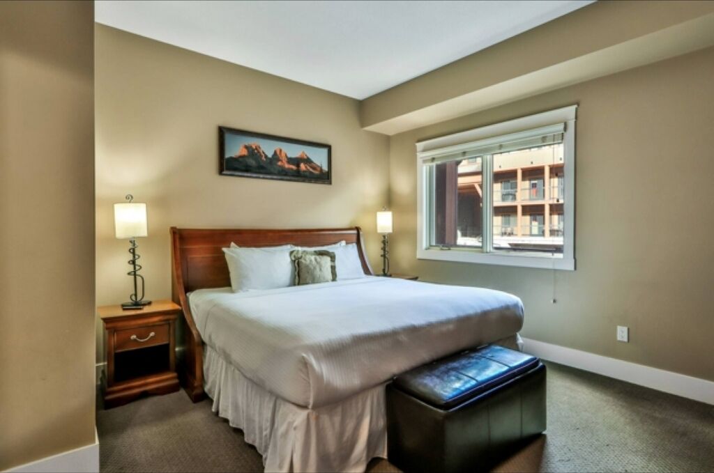 A bedroom with a large bed, two bedside lamps, a nightstand, a bench, a window with a view of buildings, and a painting on the wall.