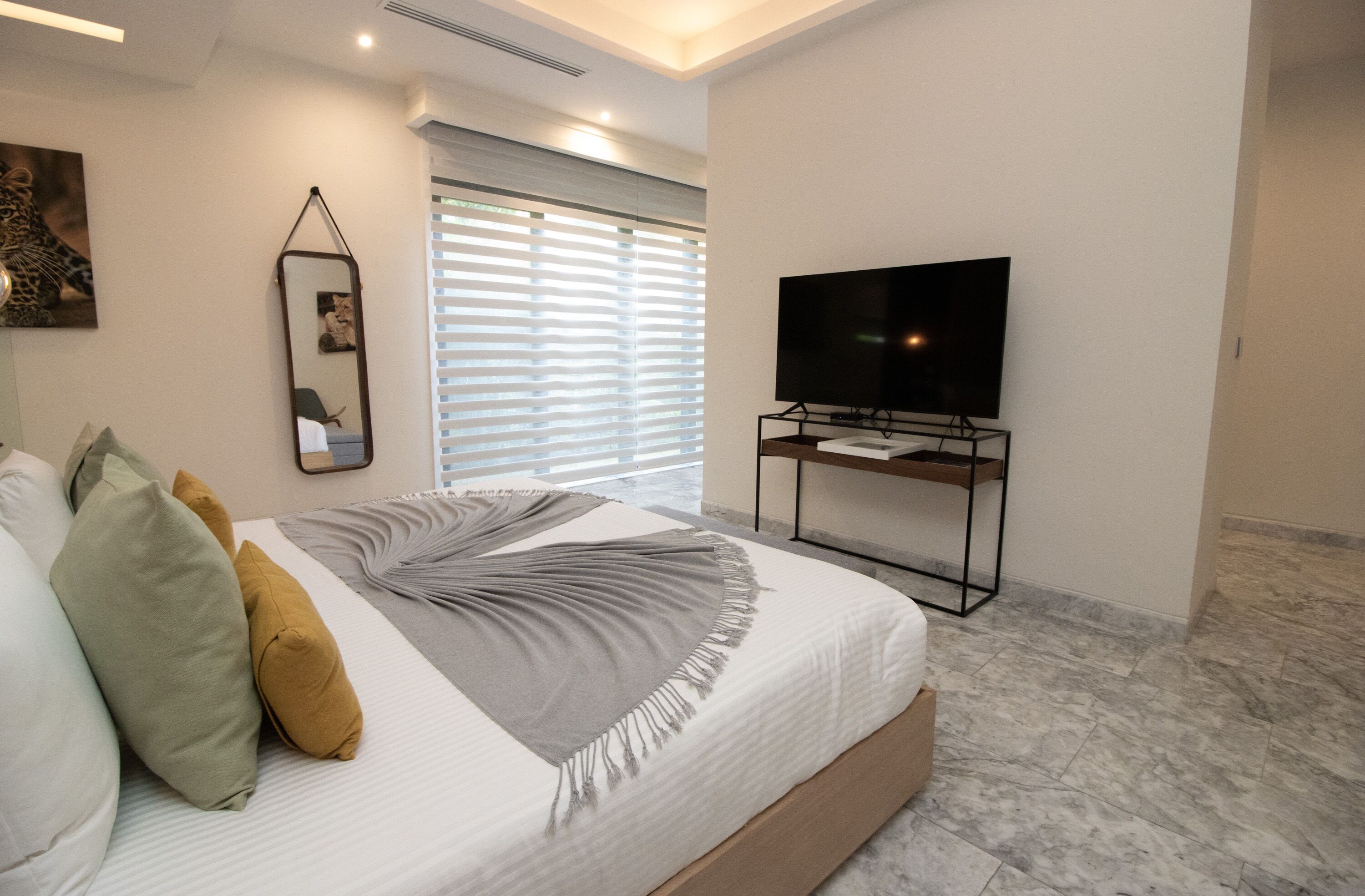 Une chambre d’hôtel moderne avec un grand lit, un téléviseur à écran plat posé sur un meuble, un miroir au mur et une fenêtre avec des stores.