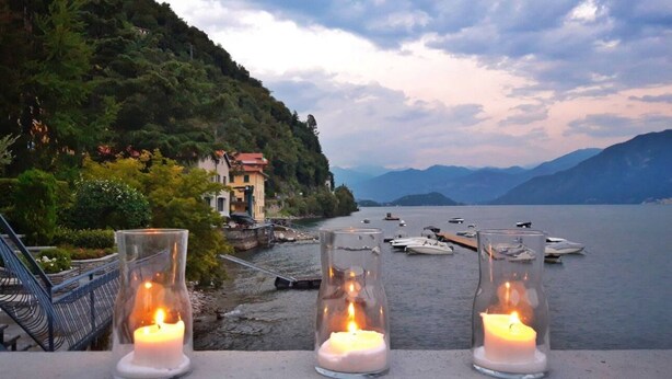 A lakeside view with three lit candles on a railing, boats on the water, and a building on the shore. - Casa Beverly 2 - My Home in Como (Argegno)