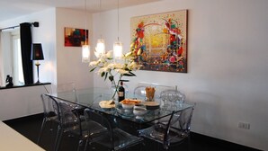 Un coin repas avec une table en verre, des chaises transparentes, un vase de fleurs et un tableau abstrait aux couleurs vives accroché au mur.