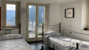 Une chambre avec deux lits, une fenêtre donnant sur les montagnes, une horloge et un tableau encadré au mur.