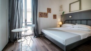 Un dormitorio con una cama, una mesita pequeña y una silla, una ventana con cortinas y cuadros en la pared.