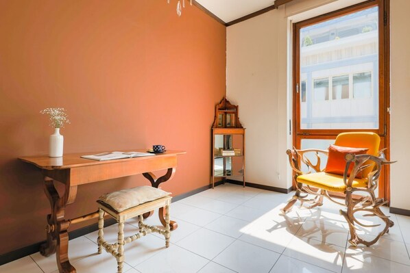 A room with a wooden desk, a small bench, a chair with a cushion, a vase with flowers, and a window with a view of buildings. - Piazza Cavour - My Home in Como (Como)