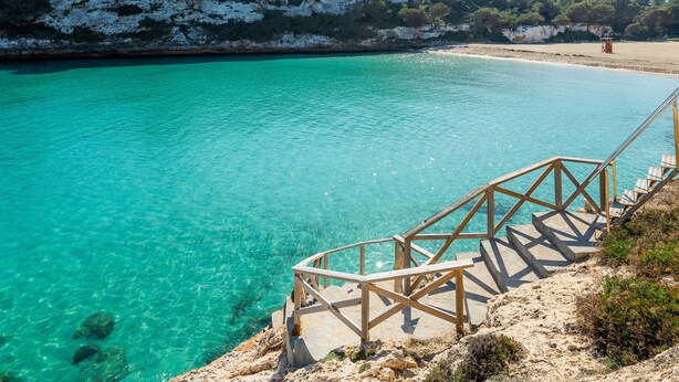 Una zona costanera amb aigua turquesa cristal·lina, una platja sorrenca i unes escales de fusta que baixen cap a l'aigua.