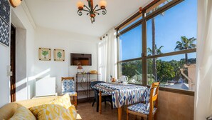 Habitació amb un llit, una taula de dinar amb cadires, un televisor i un finestra gran amb vista a palmeres.
