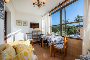 Une chambre avec un lit, une table à manger et des chaises, un téléviseur et une grande fenêtre donnant sur des palmiers.