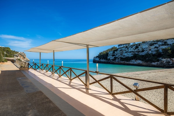 A beach with a wooden railing, a covered walkway, and a rocky cliff.