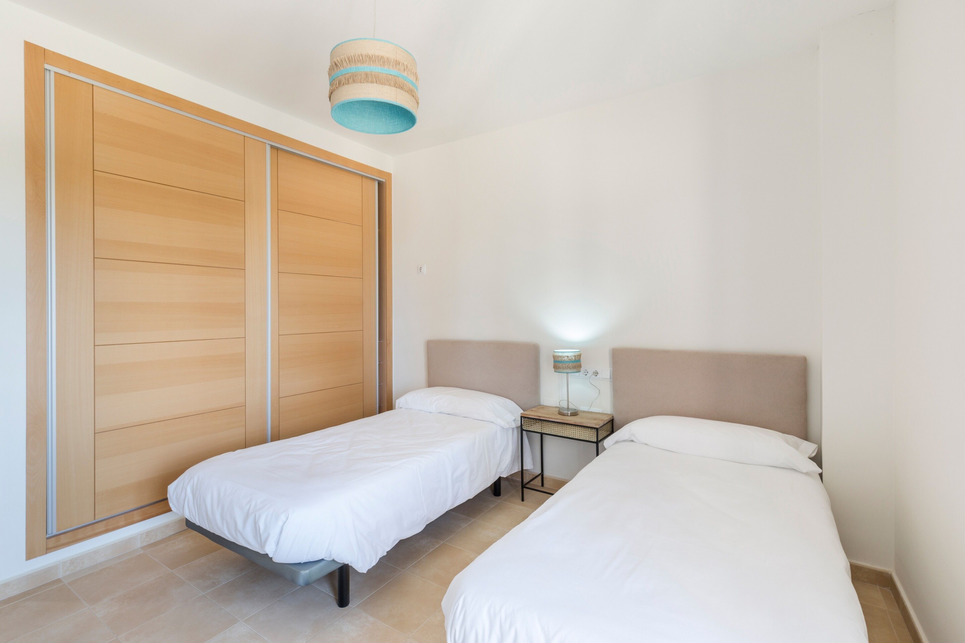 Un dormitorio con dos camas individuales, un armario de madera, una mesita de noche con lámpara y una lámpara colgante.