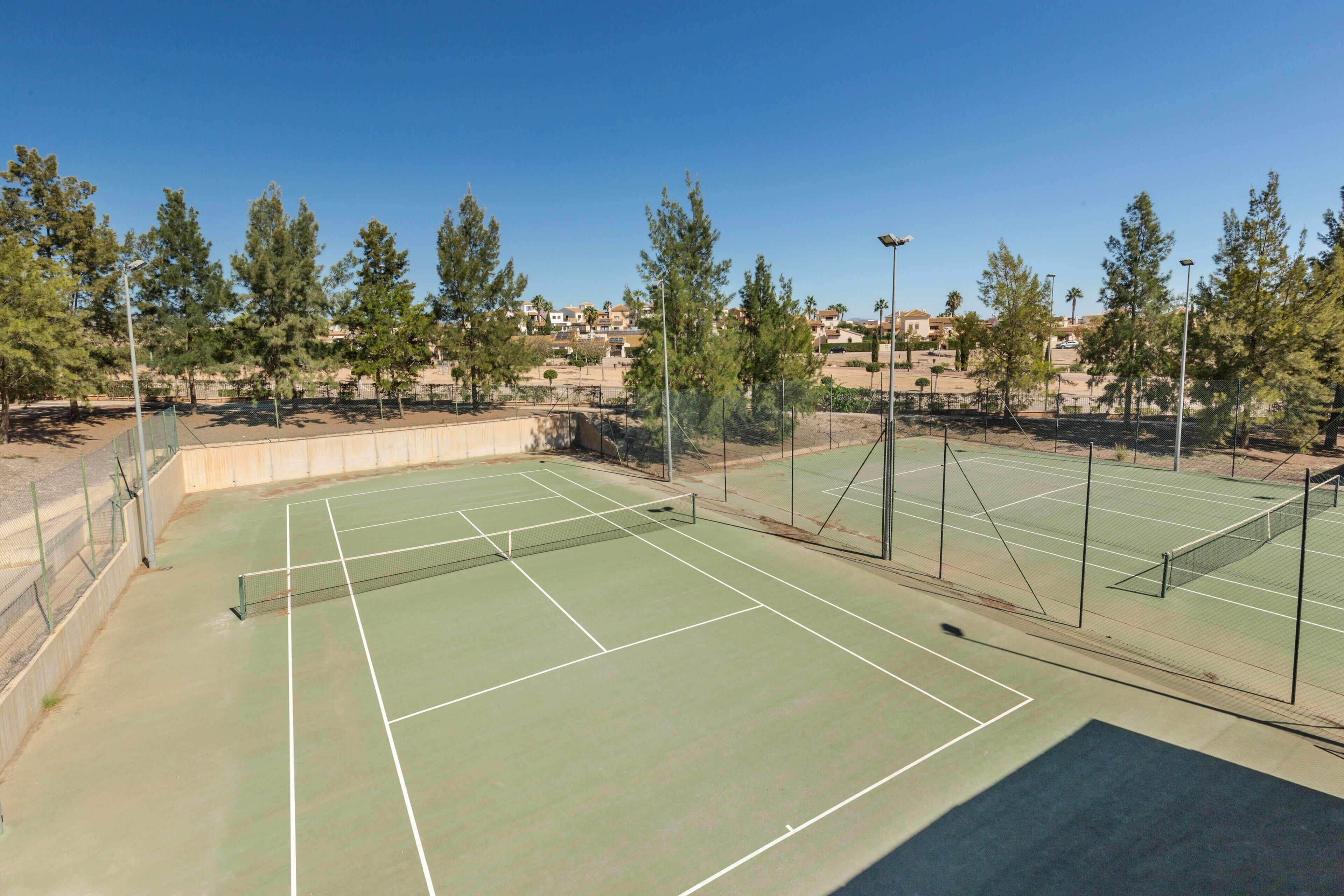 Un court de tennis en plein air, doté d’un filet, entouré d’arbres et d’une clôture.