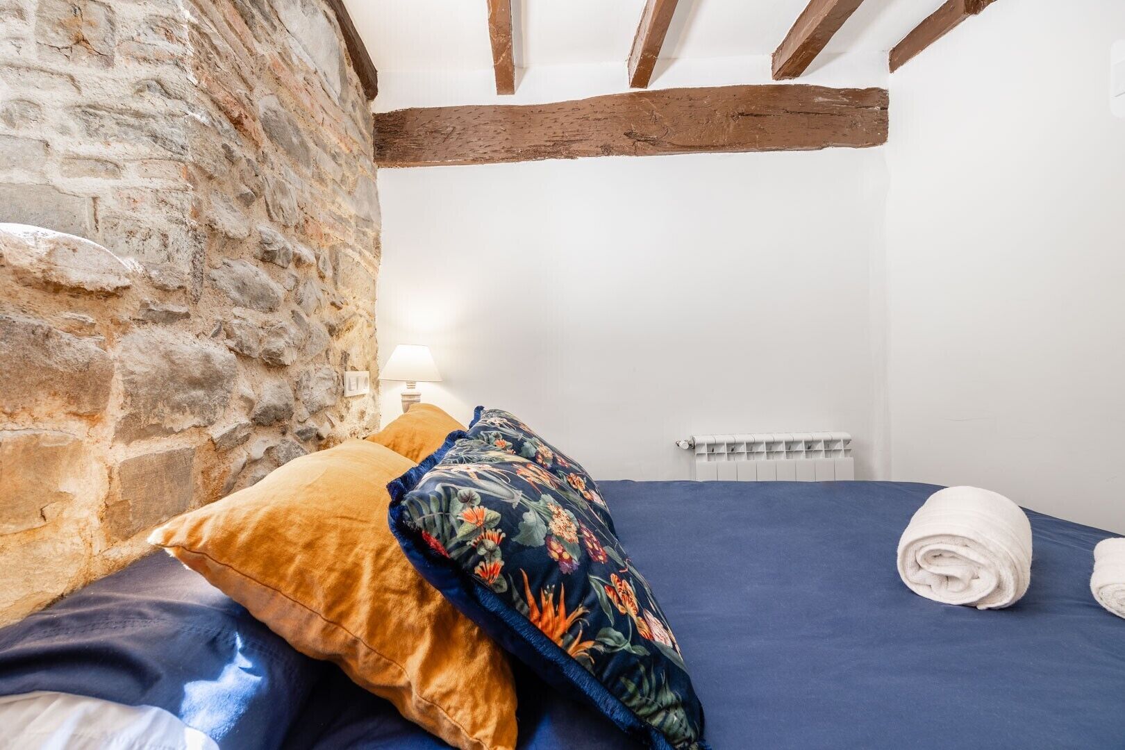 A bedroom with a stone wall, wooden beams, a bed with blue bedding, a floral pillow, and a rolled towel.
