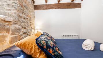 A bedroom with a stone wall, wooden beams, a bed with blue bedding, a floral pillow, and a rolled towel.