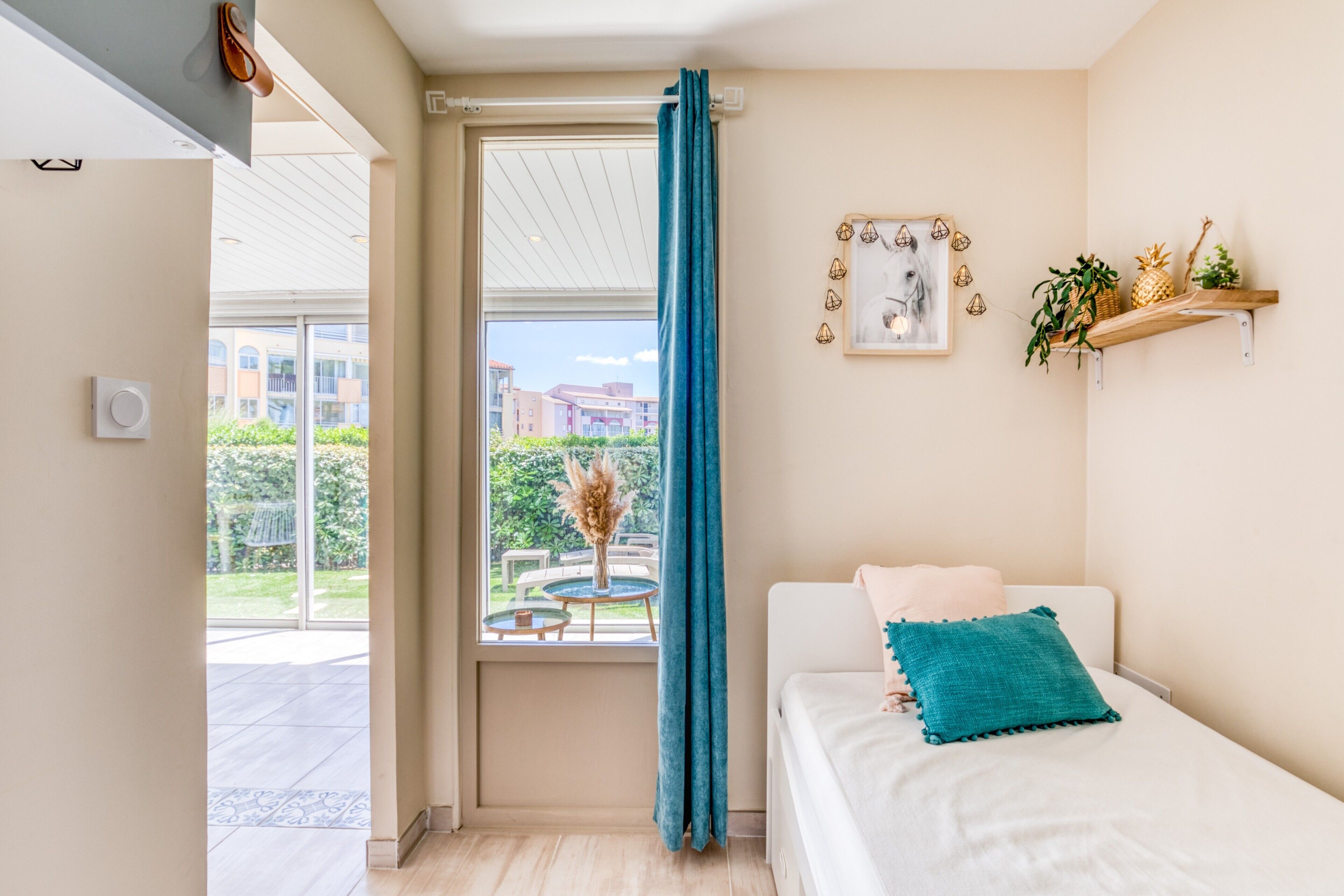 A bedroom with a bed, a window with a curtain, a wall-mounted shelf with plants, and a view of the outside.