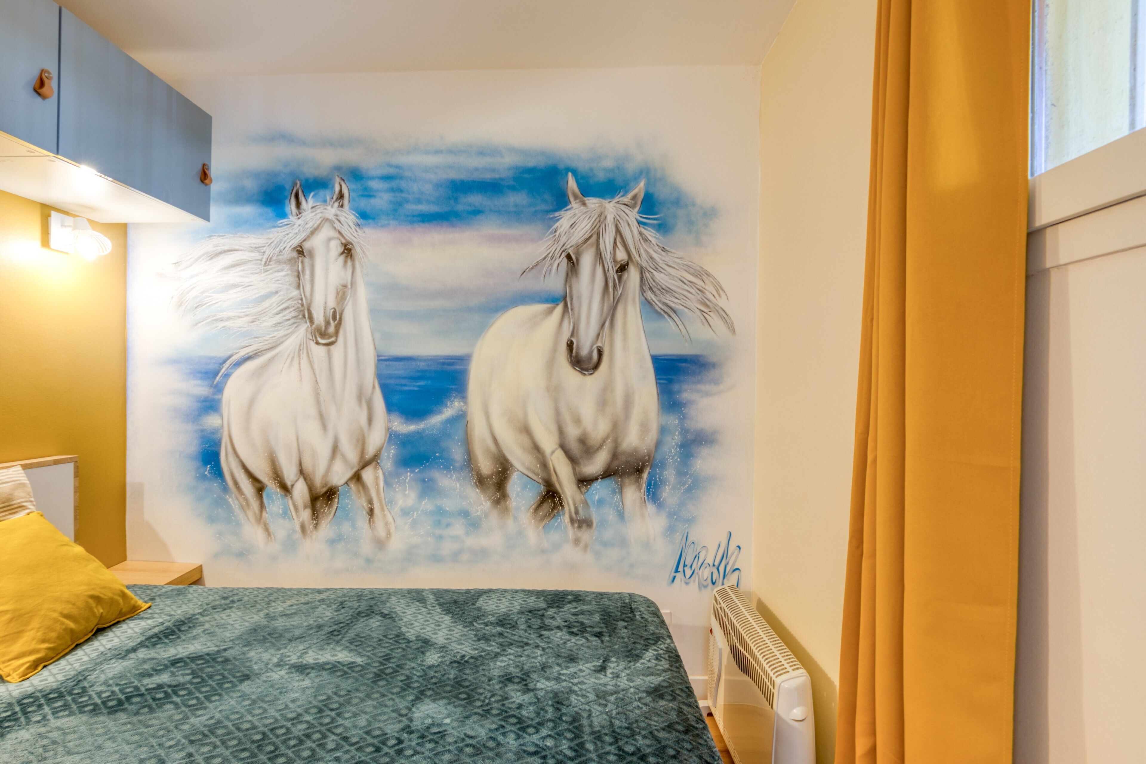 A bedroom with a bed, a nightstand, a lamp, a radiator, and a wall mural of two horses.