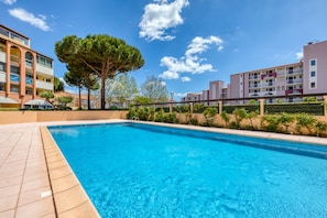 Une piscine dotée d’une terrasse carrelée, entourée de bâtiments avec des balcons et d’un grand arbre.