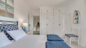 Un dormitorio con una cama, almohadas, un banco, una mesita de noche, una lámpara, un armario y un espejo.