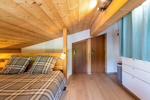 A bedroom with a wooden ceiling, a bed with plaid bedding, a bedside lamp, and a door leading to another room.