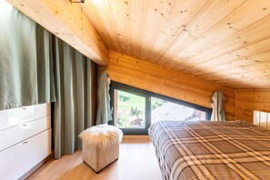 Une chambre avec un plafond en bois, un lit recouvert d’une couverture à carreaux, une commode blanche et une fenêtre avec des rideaux.