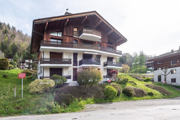 A multi-story wooden building with balconies, surrounded by greenery and hills.