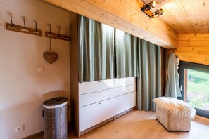 Une pièce avec un plafond en bois, une grande fenêtre avec des rideaux, un meuble blanc avec des tiroirs, une poubelle ronde et un pouf moelleux.