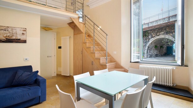 A modern living room with a blue sofa, a glass table, white chairs, a wooden staircase, and a large window with a view of a stone bridge.