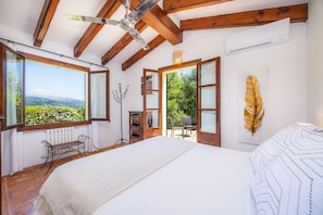 A bedroom with a large bed, a ceiling fan, wooden beams, a window with a view, and a decorative feather on the wall.