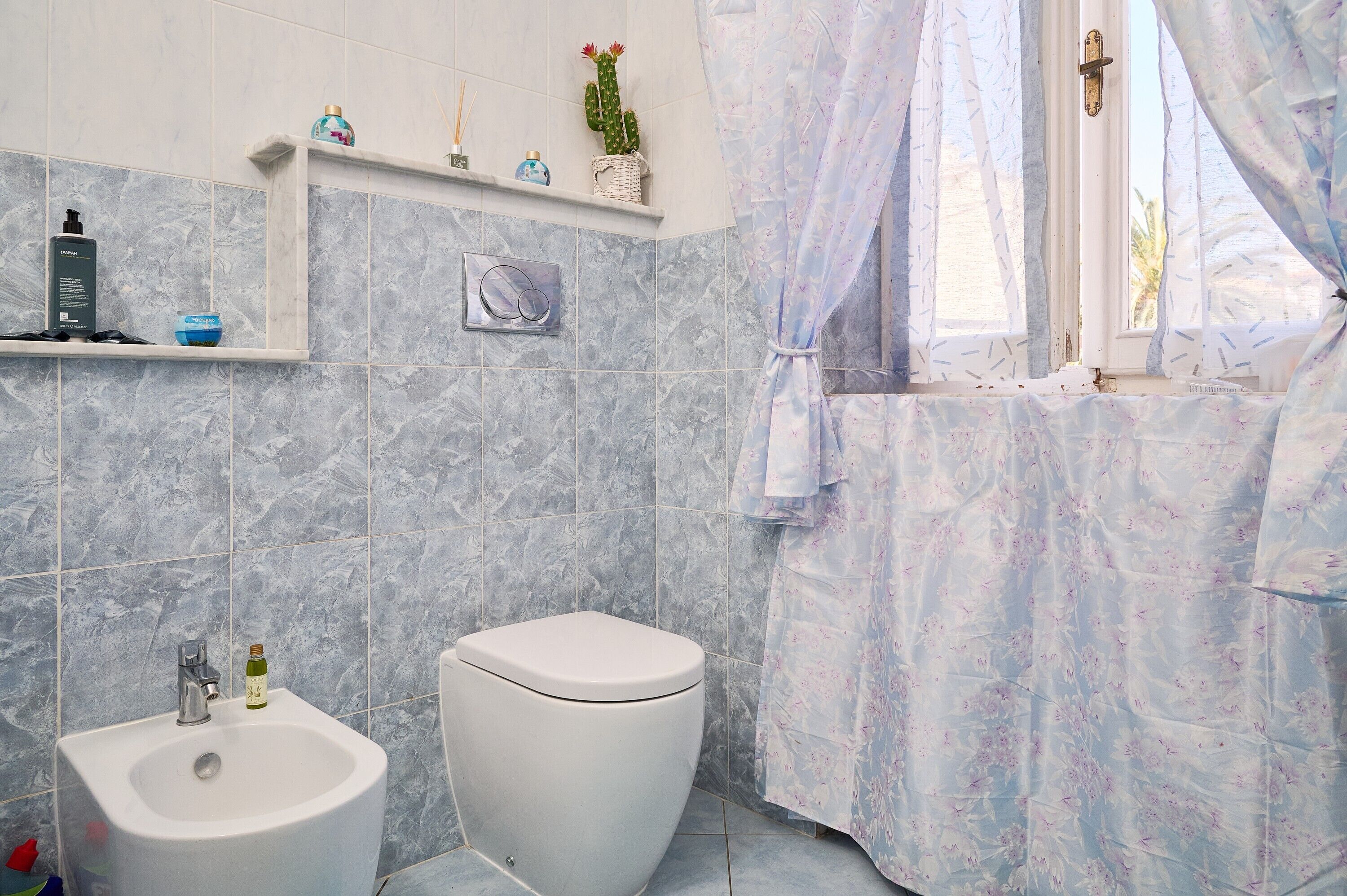 Ein Badezimmer mit Toilette, Waschbecken, Regalen mit Pflegeprodukten, einem Fenster mit Vorhängen und einem Kaktus auf dem Regal.