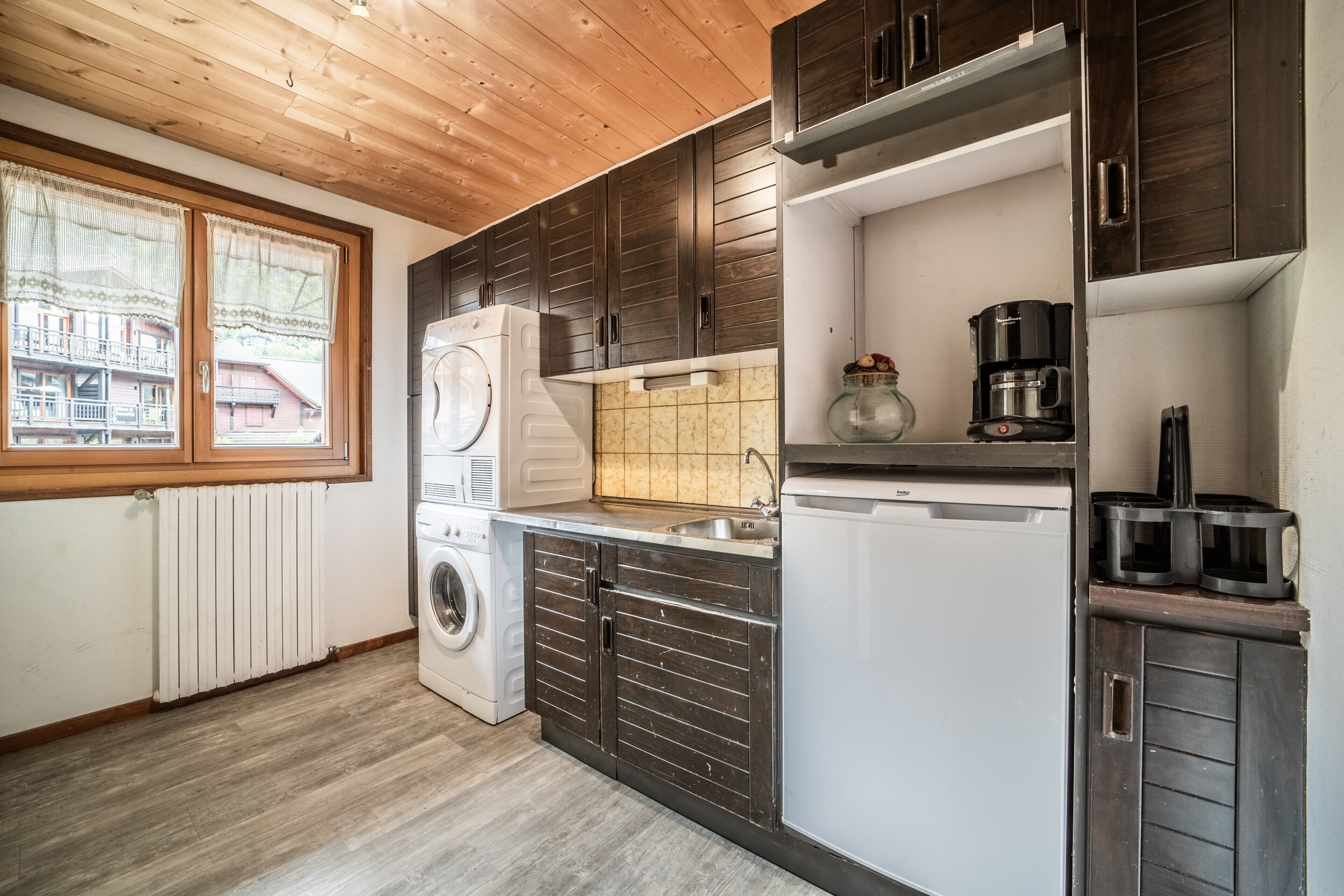 A compact kitchen with wooden cabinets, a washing machine, a coffee maker, and a view of a building through a window.