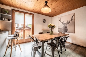 Un coin repas avec une table en bois, des chaises en métal, un vase de fleurs, une horloge au mur et un grand tableau représentant un cerf.
