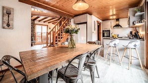 Een houten eettafel met stoelen, een bloevas, een keuken met een koelkast en een trap.
