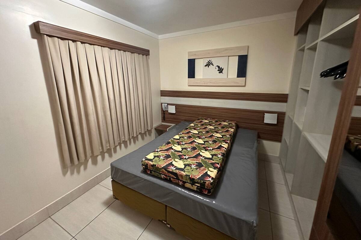 Un lit simple avec une couvre-lit à motifs, un téléphone et une œuvre d’art accrochée au mur, dans une chambre dotée d’une tête de lit en bois et d’une fenêtre avec des rideaux.