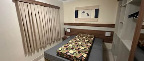 Un lit simple avec une couvre-lit à motifs, un téléphone et une œuvre d’art accrochée au mur, dans une chambre dotée d’une tête de lit en bois et d’une fenêtre avec des rideaux.