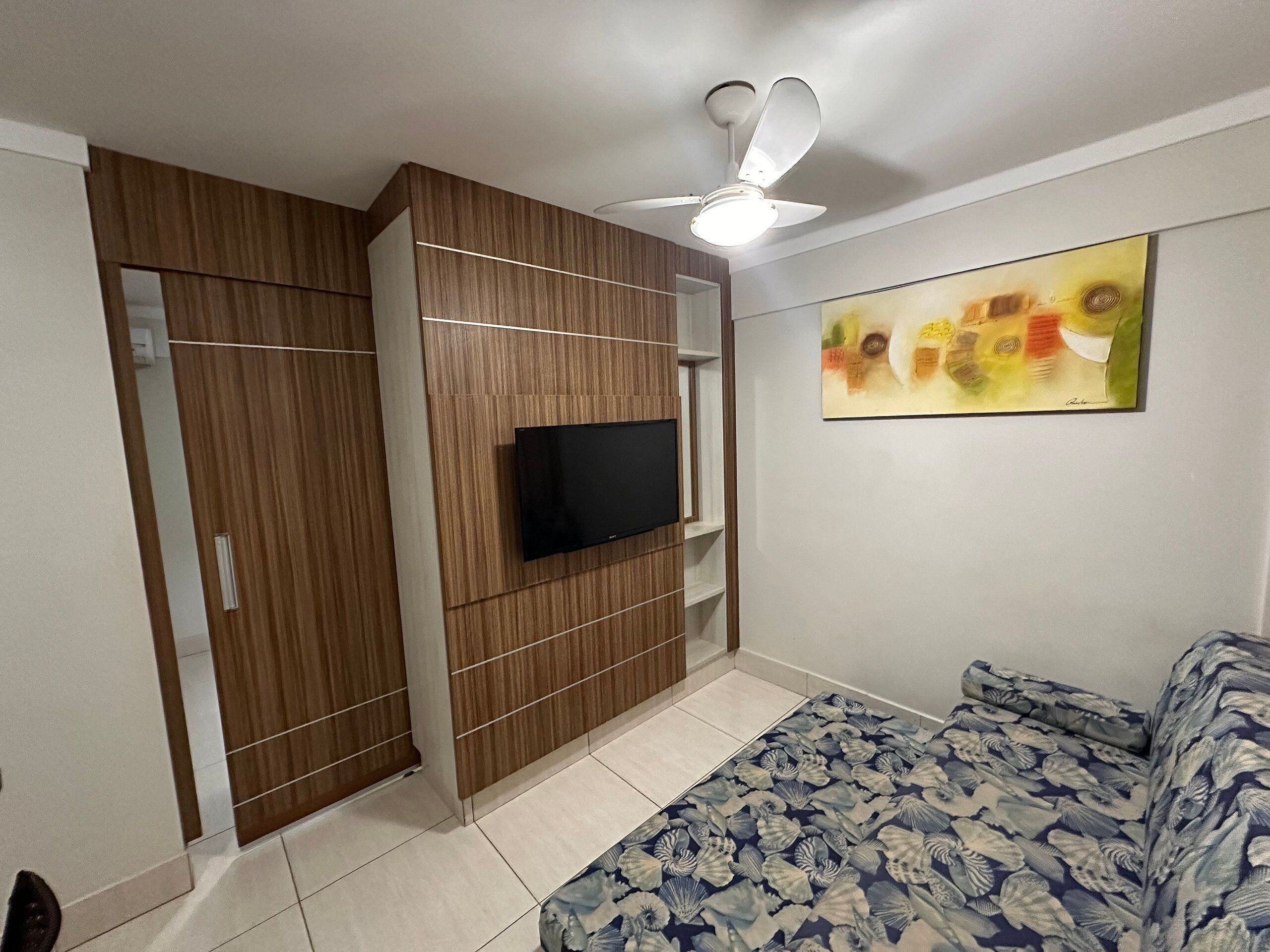 A room with a TV mounted on a wooden panel wall, a ceiling fan, and a bed with a floral-patterned bedspread.
