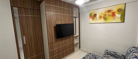 A room with a TV mounted on a wooden panel wall, a ceiling fan, and a bed with a floral-patterned bedspread.