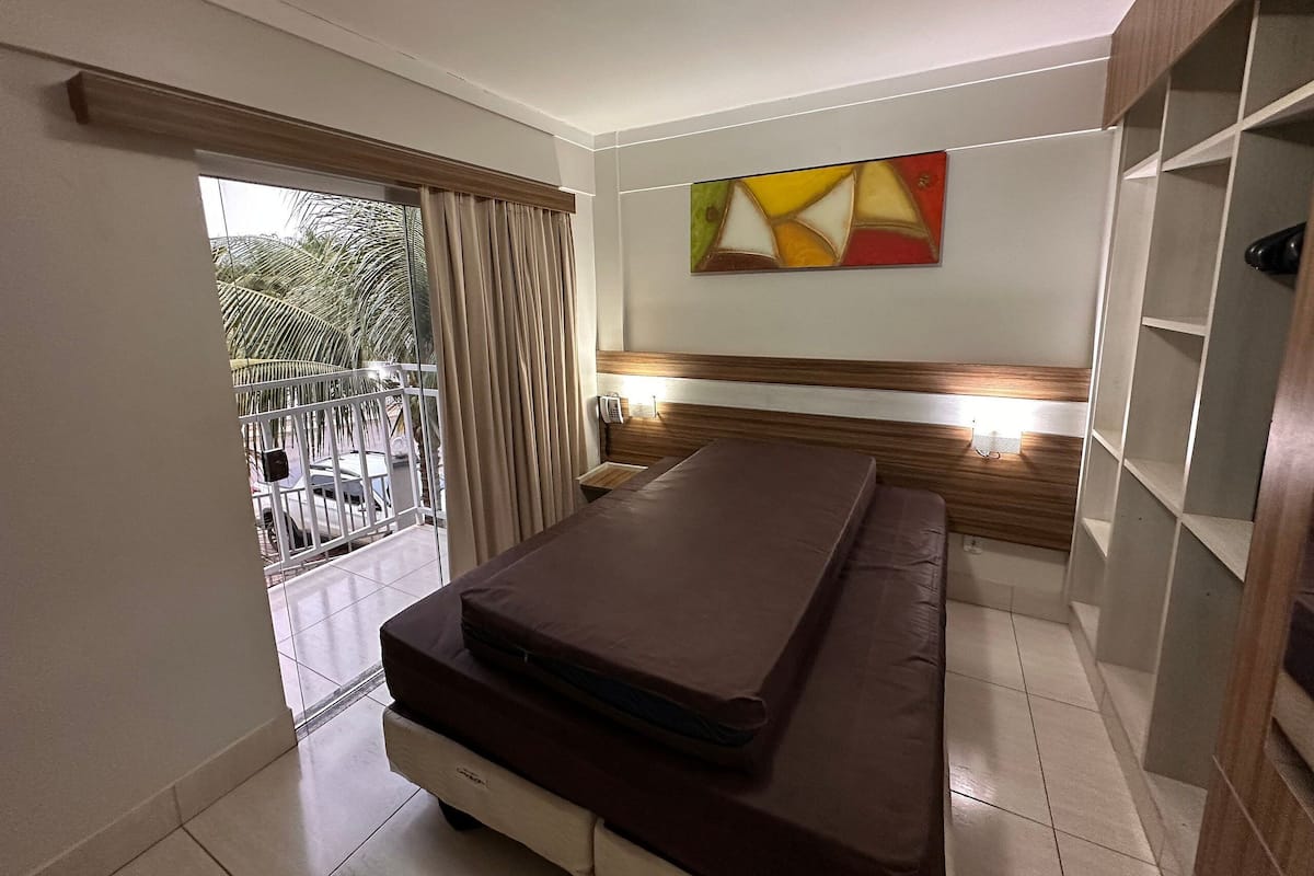 Une chambre d’hôtel avec un lit, une table de chevet, un téléphone, un tableau au mur et un balcon donnant sur des palmiers.