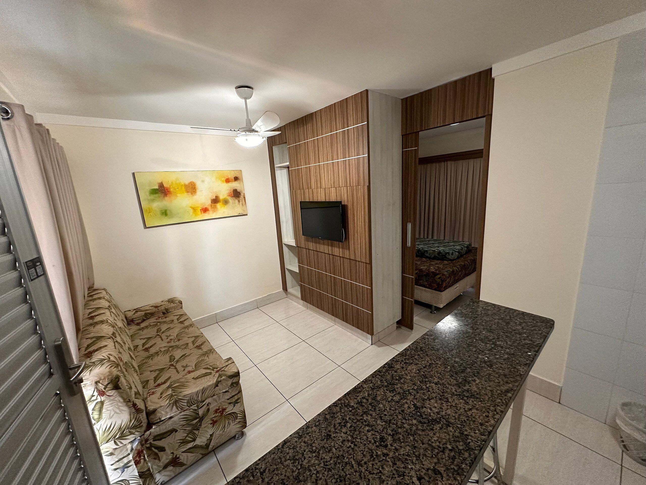 A modern living room with a sofa, a flat-screen TV, a ceiling fan, and a bed in the background.