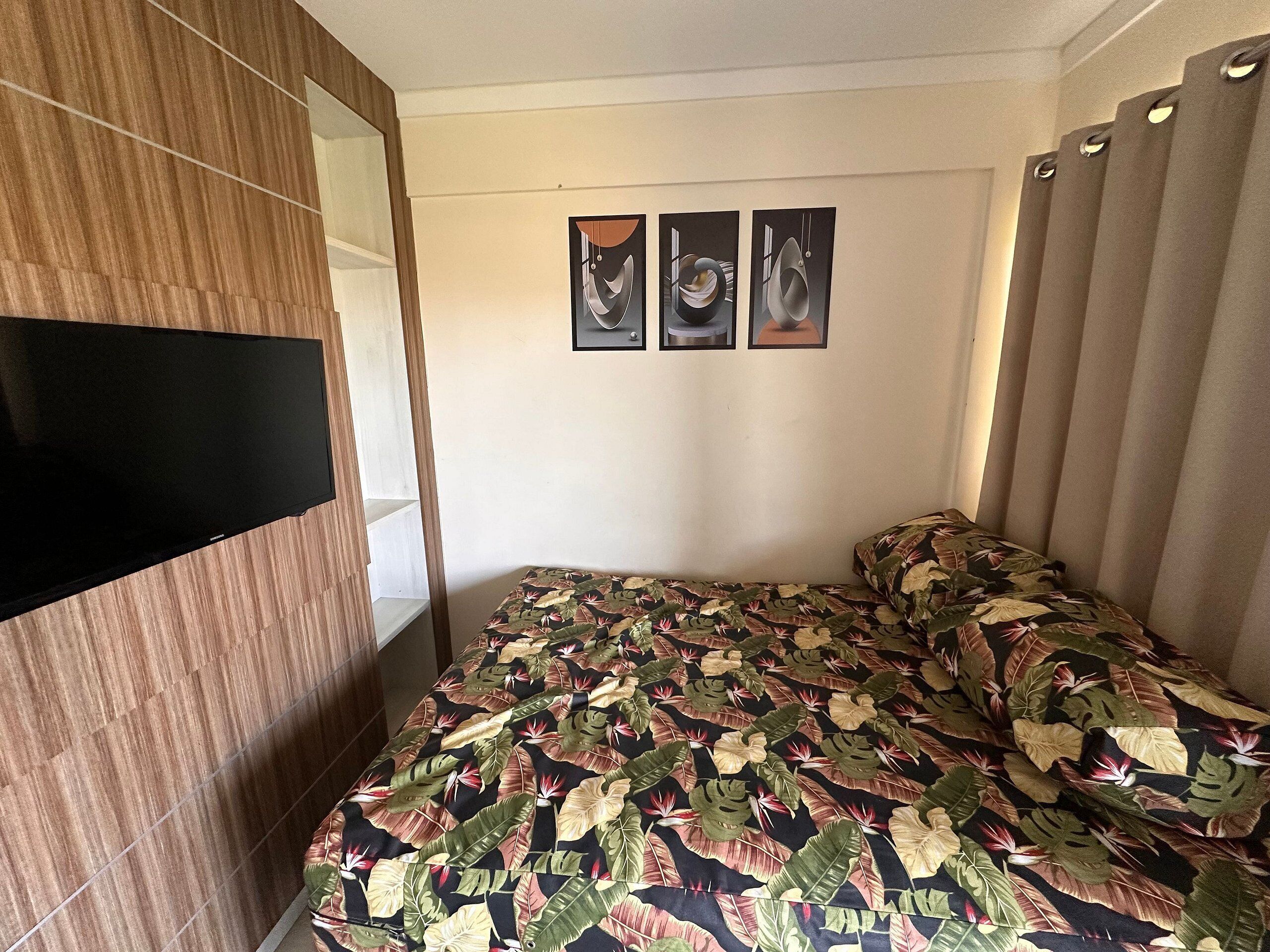 A bedroom with a bed, a TV mounted on a wooden panel, and three framed artworks on the wall.