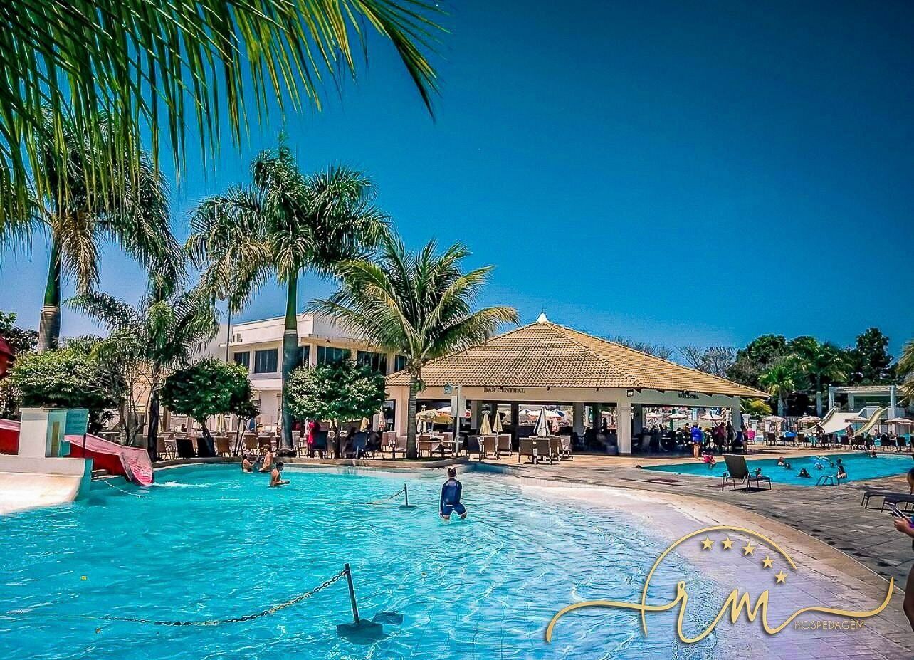 Un área de piscina con palmeras, un espacio de descanso cubierto y un edificio con tejado de azulejos.