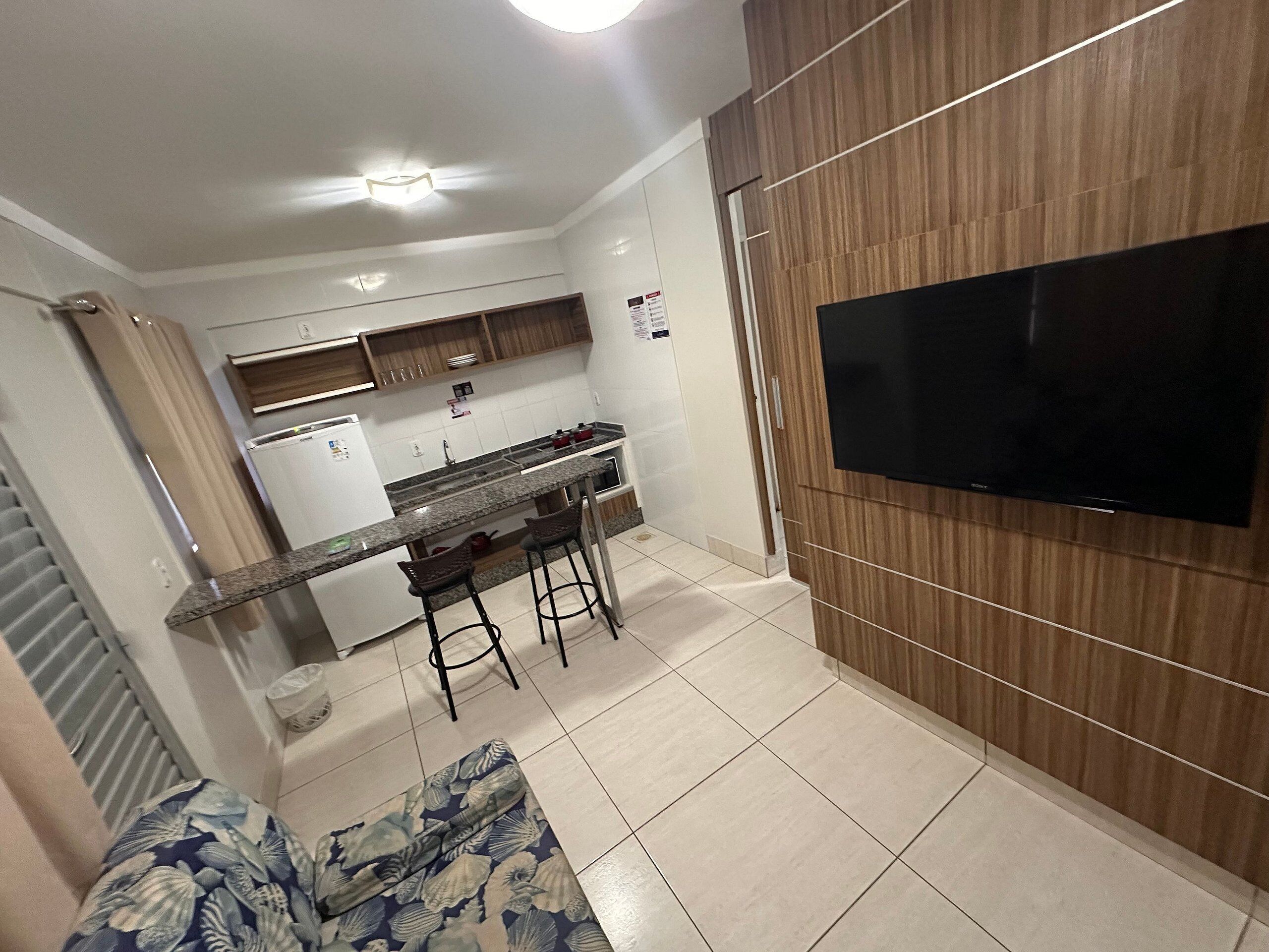 A modern kitchen with a built-in oven, a dining area with two bar stools, and a flat-screen TV mounted on the wall.