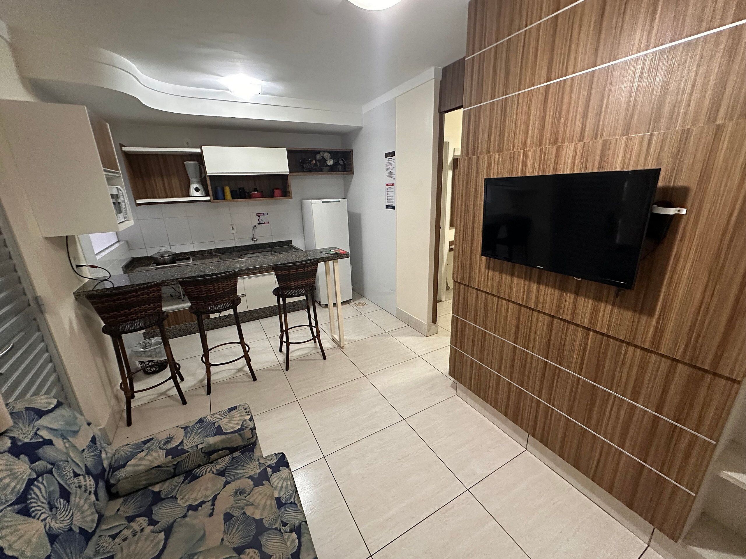 Een kleine leefruimte met een kitchenette, een eethoek met twee stoelen, een bank met bloemenprint en een aan de wand bevestigde televisie op een houten muur.