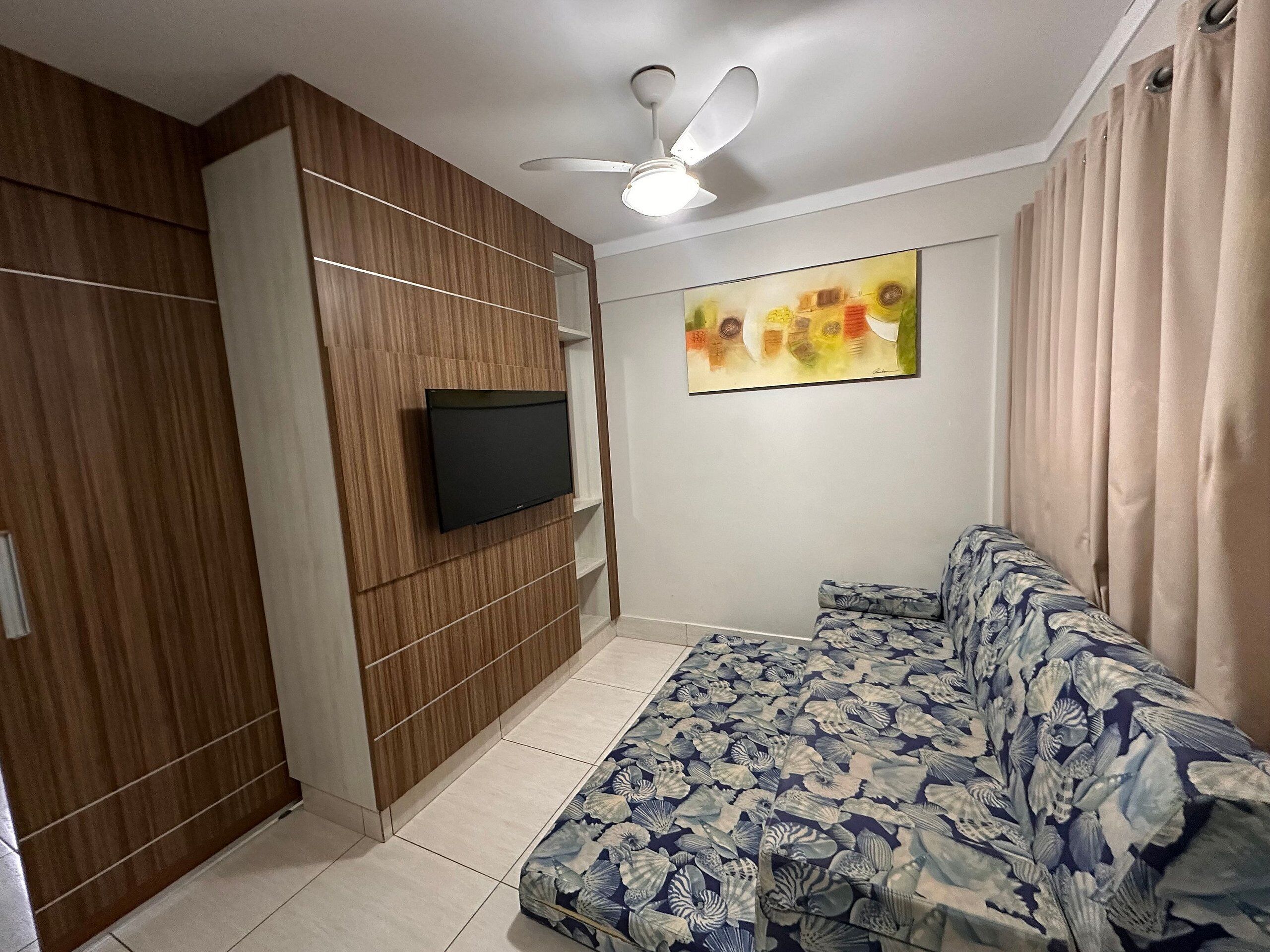 A modern living room with a flat-screen TV mounted on a wooden panel wall, a ceiling fan, a patterned sofa, and a colorful abstract painting on the wall.